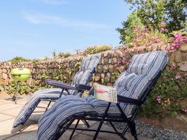 Two striped cushioned lounge chairs on a patio with a stone wall and flowers and a green barbecue grill at The Barn at Trevothen Farm in Coverack