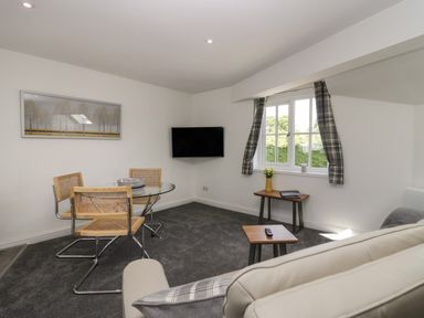 A small living room with a round glass table and three chairs a wall-mounted tv a window with plaid curtains and a small table with flowers at Betty's Loft in Hawkshead