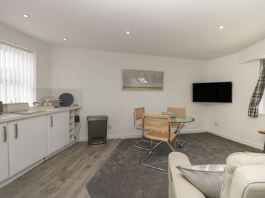 A kitchen and dining area with white cabinets a glass round table with three chairs a wall-mounted television and a trash bin at Betty's Loft in Hawkshead