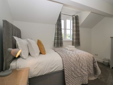A bedroom with a bed featuring white pillows and a yellow cushion a bedside table with a lamp and a window with plaid curtains at Betty's Loft in Hawkshead