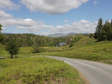 A winding road through grassy hills with trees and a lake under a partly cloudy sky at Betty's Loft in Hawkshead