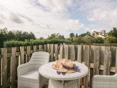 Two wicker chairs around a round wicker table with a hat and sunglasses on the table on a wooden fenced balcony with trees and buildings in the background at Betty's Loft in Hawkshead