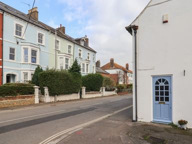 A street view with a white building with a blue door and pastel colored houses along a road at Braiders Rest in Bridport