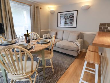 A dining area with a wooden table set with plates and wine glasses next to a beige sofa and a wooden breakfast bar with stools at Braiders Rest in Bridport