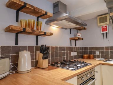 A kitchen countertop with a gas stove kettle toaster knife block and wooden shelves with glasses and a can at Braiders Rest Bridport