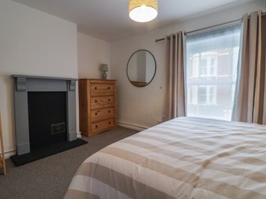 A bedroom with a bed near a window with curtains a wooden chest of drawers with a lamp a round wall mirror and a fireplace at Braiders Rest in Bridport