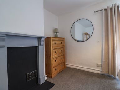 A room corner with a grey fireplace, wooden chest of drawers with a lamp, round wall mirror and beige curtains at Braiders Rest in Bridport