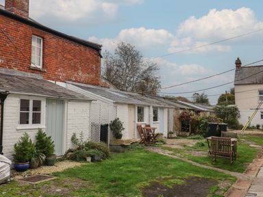 An outdoor area with white brick cottages wooden chairs and tables potted plants and a grassy yard at Braiders Rest in Bridport