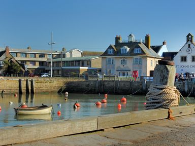 A small boat on water with red buoys near a dock with buildings and St Johns Church in the background at Braiders Rest in Bridport