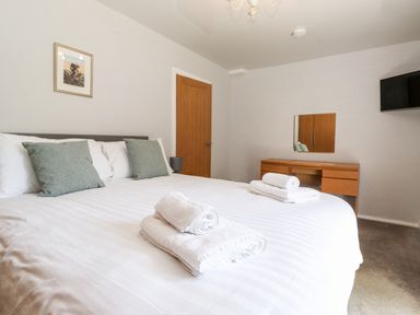A bedroom with a double bed with white linens and grey pillows two sets of folded white towels on the bed a wooden door a wooden desk with a mirror and a wall-mounted television at Fell View Clappersgate near Ambleside