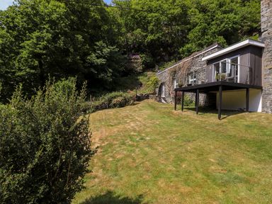 A grassy garden area with bushes and a stone house with a raised deck at Fell View in Clappersgate near Ambleside