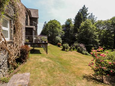 A garden with grass, bushes, and trees next to a stone house with a raised deck at Fell View in Clappersgate near Ambleside