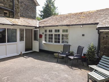An outdoor area with two black chairs a green bench a small tree near a white wall with windows and an open door at Fell View in Clappersgate near Ambleside