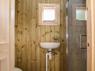 A small bathroom with wooden walls a white sink a small window and a glass shower door at The Oak Hut in Coton near Whitchurch