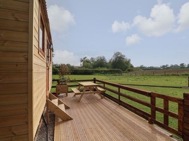 A wooden deck with a picnic table plant pot and staircase overlooking a green field at The Oak Hut in Coton near Whitchurch