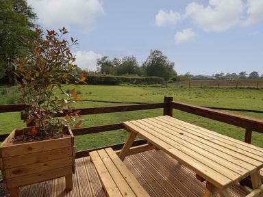 A wooden picnic table and bench on a deck with a planter and green fields in the background at The Oak Hut in Coton near Whitchurch