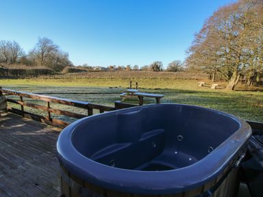 An outdoor hot tub on a wooden deck overlooking a grassy field with trees and sheep at The Oak Hut in Coton near Whitchurch