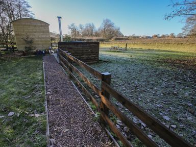 A gravel path next to a wooden fence and a wooden structure with a chimney in a grassy field with a picnic table at The Oak Hut in Coton near Whitchurch