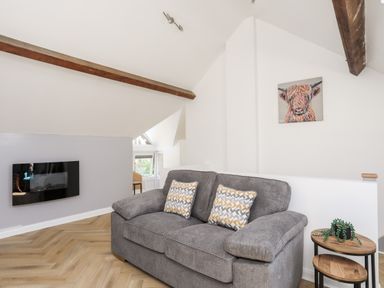 A living room with a gray sofa two patterned cushions a small wooden side table with a plant a wall-mounted TV and a colorful cow painting on the wall at Stabal Bach in Criccieth