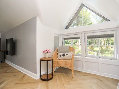 A wicker chair with a cushion and throw blanket next to a small round wooden table with flowers near triangular windows at Stabal Bach in Criccieth