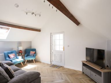 A living room with two blue chairs, a grey sofa, a small table lamp, a wall-mounted television and wooden beams on the ceiling at Stabal Bach in Criccieth