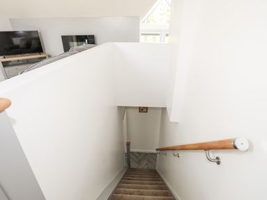 A staircase with wooden handrail leading down to a room with carpeted steps and a TV visible upstairs at Stabal Bach in Criccieth