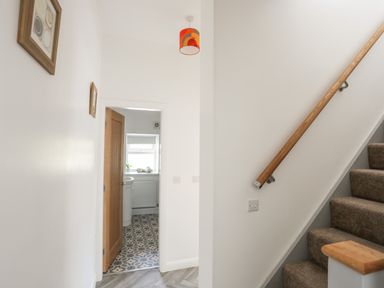 A hallway with carpeted stairs and wooden handrail leading up and an open doorway showing a bathroom with patterned floor tiles at Stabal Bach in Criccieth