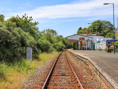 A railway track leading to a small train station platform with a white building and greenery surrounding at Stabal Bach in Criccieth