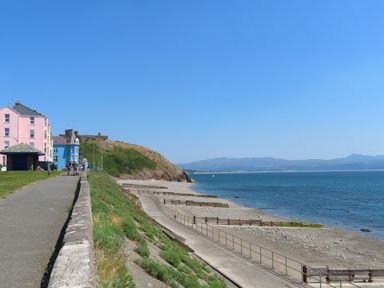 A seaside path with people walking near pastel-colored buildings and a beach with railings at Stabal Bach in Criccieth