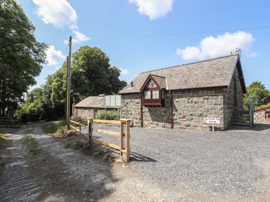 A gravel driveway and parking area next to stone buildings with trees and a wooden fence at Stabal Bach in Criccieth