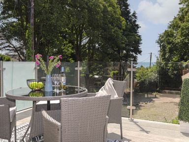 An outdoor patio with a glass table surrounded by wicker chairs and a vase with flowers at Stabal Bach in Criccieth