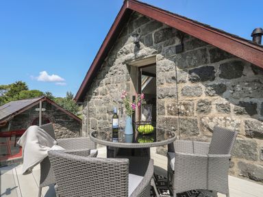 An outdoor patio with wicker chairs around a glass table holding a vase with flowers a bottle and a fruit bowl at Stabal Bach in Criccieth
