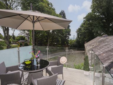 A balcony with a glass table chairs an umbrella and decorative plants overlooking trees and a gravel area at Stabal Bach in Criccieth