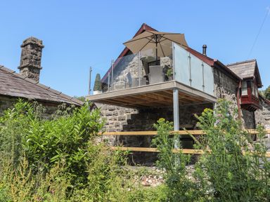 A stone house with a raised balcony and patio furniture with umbrella surrounded by plants at Stabal Bach Criccieth