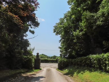 A driveway bordered by hedges and trees leading to an entrance with stone pillars by a road at Stabal Bach in Criccieth
