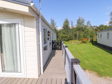 An exterior view of a beige mobile home with a wooden deck and railing overlooking a grassy area with trees at 41 Grandmas Cottage South Lakeland Leisure Village near Carnforth