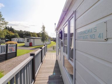 A wooden walkway along a white cottage with a sign reading Grandma's Cottage No 41 and neighboring cottages in a grassy area at South Lakeland Leisure Village near Carnforth