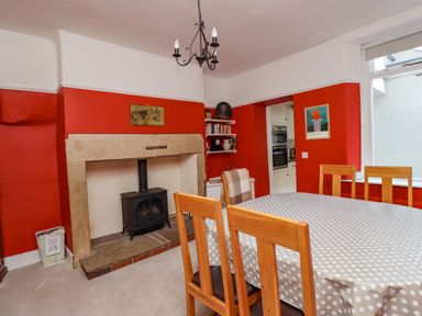 A dining room with a polka dot tablecloth on a table wooden chairs a black stove in a stone fireplace red walls and a window at Victoria House in Alnwick