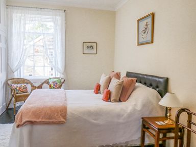 A bedroom with a bed and pillows a side table with a lamp wicker chairs by a window with lace curtains at Victoria House in Alnwick