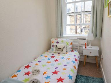 A small bedroom with a single bed covered in star patterned bedding next to a window with curtains and a white nightstand with a lamp and an orange object at Victoria House in Alnwick