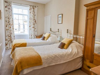 A bedroom with two single beds yellow throws pillows a wooden wardrobe and a window with patterned curtains at Victoria House in Alnwick
