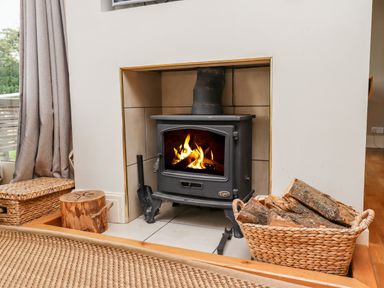 A wood burning stove with a fire burning inside a tiled fireplace with a basket of firewood nearby at Cullernose Cottage Newton-by-the-Sea near Embleton