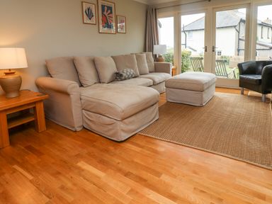 A living room with a beige sofa and ottoman a wooden side table with a lamp and framed prints on the wall at Cullernose Cottage Newton-by-the-Sea near Embleton