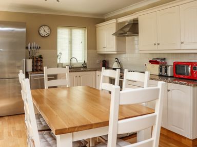 A kitchen with a wooden dining table and white chairs near a window with blinds and kitchen appliances at Cullernose Cottage in Newton-by-the-Sea near Embleton