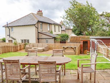 An outdoor patio with wooden table and chairs a garden with a soccer goal a shed and houses in the background at Cullernose Cottage in Newton-by-the-Sea near Embleton