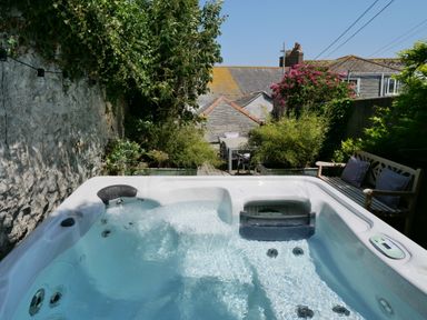 An outdoor hot tub with water surrounded by plants a wooden bench and a table with chairs in the background at Waves End in St Ives