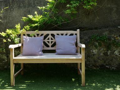 A wooden bench with two cushions in a garden with a stone wall and green plants at Waves End in St Ives