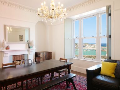 A dining room with a large wooden table, chairs, a chandelier, a fireplace, and a window overlooking a harbor at Waves End in St Ives