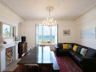 A dining room with a wooden table and chairs a black leather sofa and a window with a sea view at Waves End in St Ives