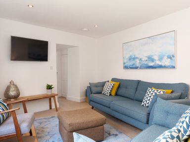 A living room with blue sofas a brown ottoman a wooden chair a wall-mounted television and a blue and white painting at Waves End in St Ives